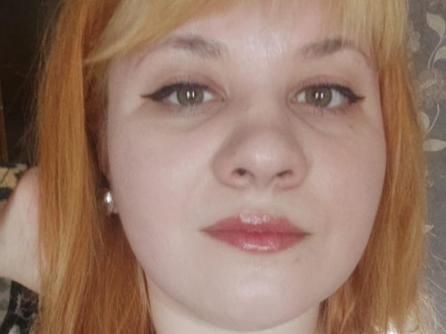 AlisaMi has pale skin, red-orange hair, and wears black eyeliner. Her pink lips are slightly glossy. She has small pearl earrings and a neutral expression.