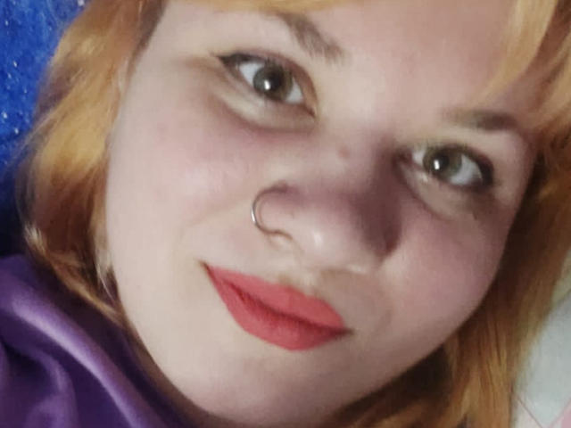 AlisaMi's face with pale skin, red lipstick, and a nose ring. She has blonde hair and green eyes, smiling slightly. She's wearing a purple shirt.