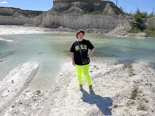 AlisaMi stands in bright neon yellow pants, black t-shirt, and gray shoes, with a small black purse, on white gravel by a clear blue-green waterhole, under sunny skies.