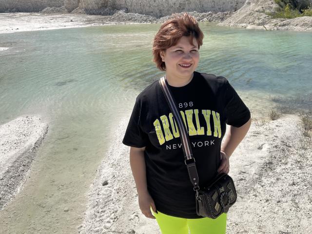 AlisaMi with short brown hair, smiling, wearing a black "Brooklyn New York" t-shirt and neon yellow pants, black camera bag, standing in a rocky, turquoise water area.