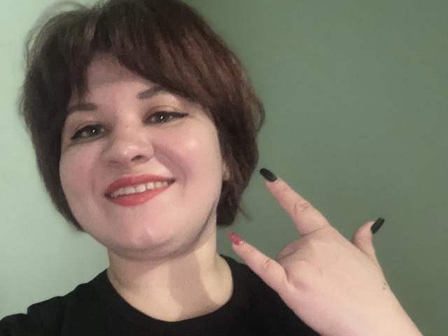 AlisaMi has short brown hair, red lipstick, black nails, and is smiling with her finger up, wearing a black shirt. Her face is slightly chubby.