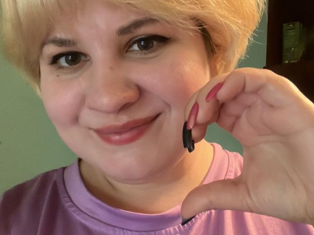 AlisaMi with short blonde hair, smiling, holding a black eyeliner near her right eye, wearing a purple shirt, pink nails, and a nose ring.