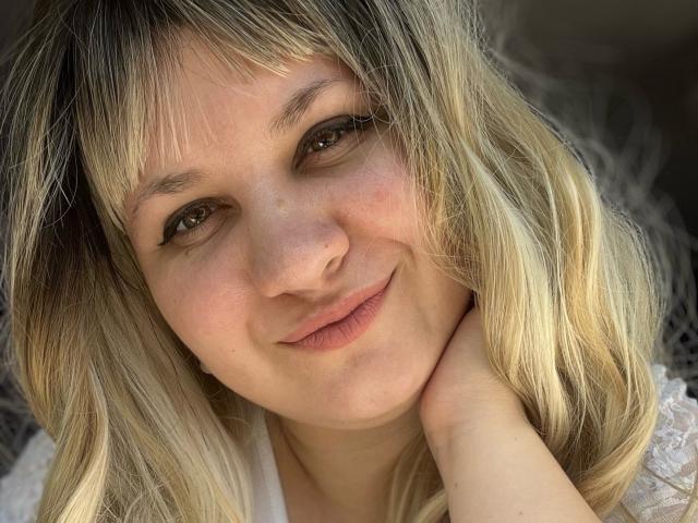AlisaMi has blonde, wavy hair with bangs, light skin, and a gentle smile. She's touching her neck, showing a relaxed, cute face with soft brown eyes.