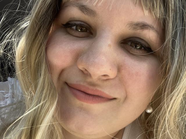 AlisaMi's face with blonde hair, light skin, brown eyes, subtle smile, and pearl earring, showing small nose and natural, slightly parted pink lips. Her makeup includes black eyeliner.