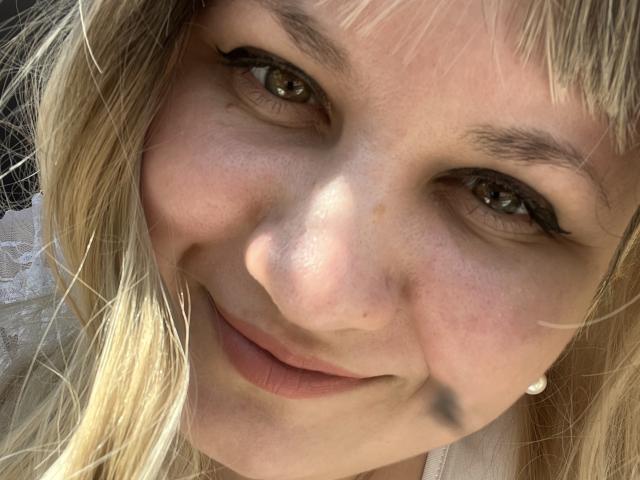 AlisaMi's face, showing a smile with light blonde hair, green eyes, black eyeliner, small nose, light freckles, and a small black mark on her right cheek.