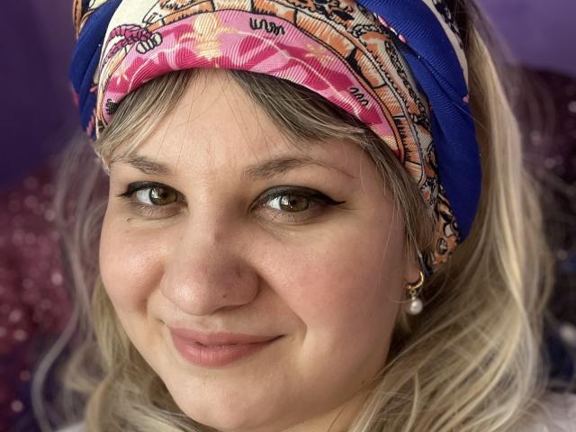 AlisaMi's face with a pink and blue headscarf, brown eyes, black eyeliner, small nose, and a slight smile. She has blonde hair, pearl earrings, and a cute look.