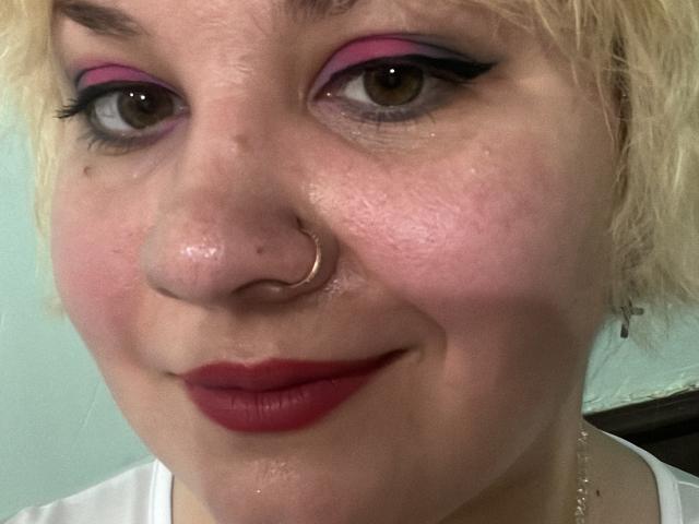 AlisaMi's face with pink eyeshadow, black eyeliner, red lipstick, nose ring, and blonde curly hair, showing a slight smile. Her skin is slightly shiny, and she's wearing a white shirt.