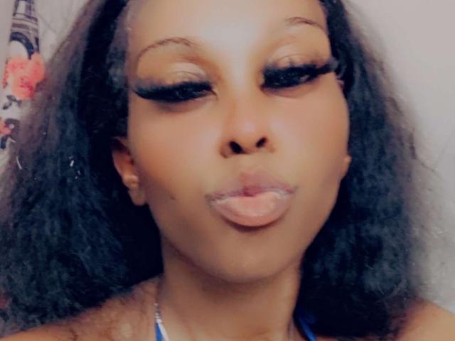 BellaStella69 has dark brown curly hair, thick black eyelashes, and puffed pink lips. She's making a duck face with her nose slightly up. Her skin is a warm brown.
