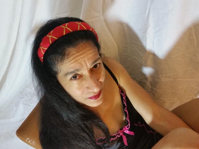 AnalCab, an older woman with long black hair, red headband, and black lingerie with pink trim, looking up with red lipstick and a serious expression.