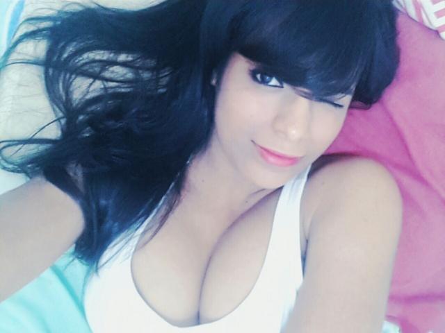 Caffein has black hair, winks, and wears a white tank top showing her big tits. She's lying on a pink pillow with a light skin tone and pink lips.