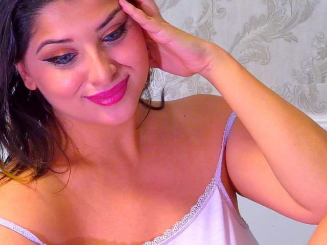 ScarletOra with pink lipstick, dark eyebrows, and white spaghetti-strap top, touching her head with one hand, smiling at the camera, showing her tits and a cute face.