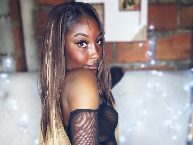 TanishaX, avec sa peau brun foncé, ses longs cheveux tressés, son haut en maille noire qui dévoile ses seins, regardant en arrière avec un léger sourire, un anneau au nez et de doux yeux marron..