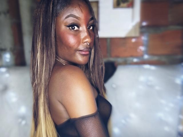 TanishaX hat eine dunkelbraune Haut, langes, glattes, braunes Haar mit blonden Strähnen und trägt ein schwarzes, durchsichtiges Oberteil, das ihre nackten Schultern und kleine, feste, pralle Brüste zeigt. Sie schaut zurück..