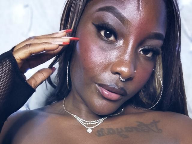 TanishaX with dark skin, red nails, hoop earrings, nose ring, and a necklace, showing a tattoo on her chest, looking at the camera with a subtle smile.