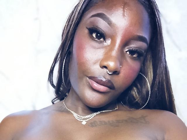 TanishaX a la peau brun foncé, de longs cheveux lisses, des boucles d'oreilles, un anneau au nez et un collier. Elle regarde l'appareil photo avec un léger sourire, et on peut apercevoir une petite cicatrice..
