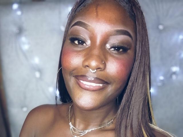 TanishaX a la peau brun foncé, de longs cheveux bruns et lisses, un anneau au nez et un sourire avec des lèvres roses et brillantes. Elle porte un collier et a un maquillage discret avec du blush rose..