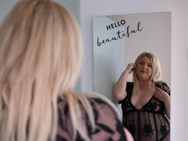 BlondineEmanuellaGrey with blonde hair, wearing a sheer black lace nightie, stands in front of a mirror, looking at her reflection, touching her hair, showing her tits.