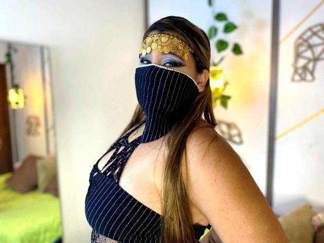 SalmaAzaman with long brown hair, wearing a black pinstripe mask, gold headpiece, and black lace-up top, showing her tits, green eyes, and a confident smile.
