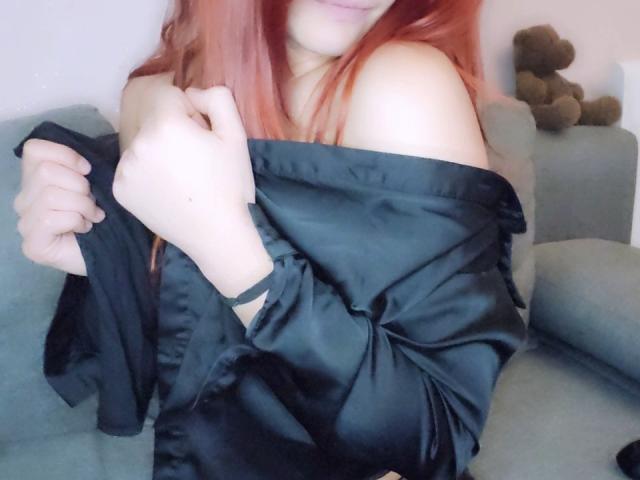 LilouLove with red hair, off-shoulder black satin robe, slightly pulling it down, exposing her bare shoulder, sitting on a gray couch, hand near her neck, smiling.