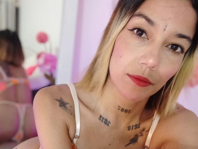 Veroo69, avec ses cheveux blonds, son rouge à lèvres rouge et ses tatouages sur la poitrine et les épaules, portant un soutien-gorge blanc, assise devant un miroir, montrant son reflet..