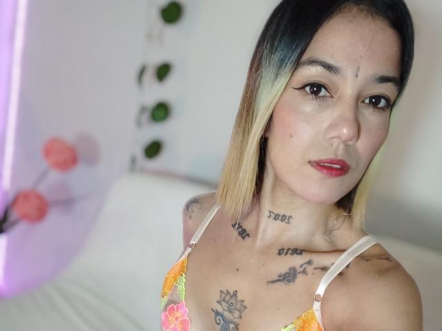 Veroo69 with blonde-to-black ombre hair, wearing a floral bra, showing tattoos on chest and neck, red lipstick, and a confident look, in a brightly lit room.