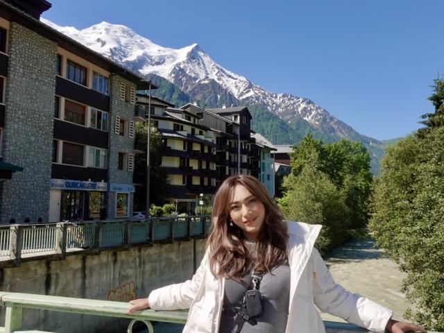 GorgeousQueen with brown hair, wearing a white jacket and grey top, stands by a green railing, snow-capped mountains in the background, clear blue sky, green trees on the right.