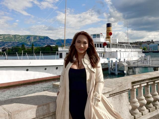 GorgeousQueen with long brown hair, wearing a cream coat over a black dress, standing by a white boat with a yellow funnel, holding her coat, smiling.