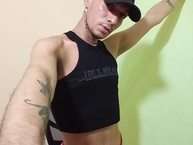 NathanTwentyTwo, wearing a black cap and sleeveless crop top, showing a tattooed arm, with a slight smirk, and red underwear, against a light green wall.