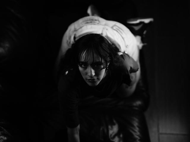 CamilaMarie is on all fours, doggy-style, with a white shirted guy behind her, his cock partially visible, in a black and white shot, looking up with intense eyes.