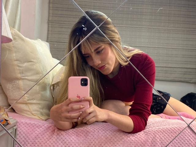 LivelyVanderWood, blonde, in red sweater, lying on pink bed, looking at pink phone, mirror reflection, hands on phone, cute, relaxed, bedroom, soft lighting.