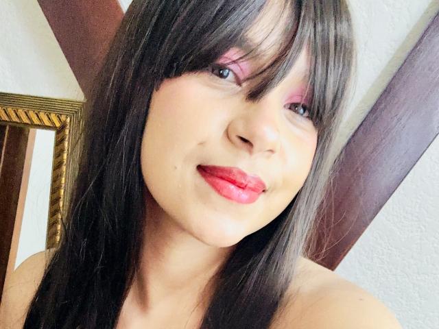 AngelinePetti tiene el cabello negro y liso, sombra de ojos rosa y un lápiz labial rojo brillante, sonriendo ligeramente con una expresión linda y suave, mostrando un pequeño grano en su mejilla derecha..