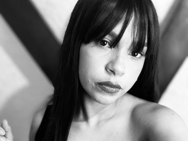 Fotografía en blanco y negro de AngelinePetti con cabello oscuro y lacio, flequillo, labios brillantes y saliva visible en su barbilla, mirando hacia arriba con una expresión ligeramente fruncida, sin ropa en la parte superior del cuerpo..