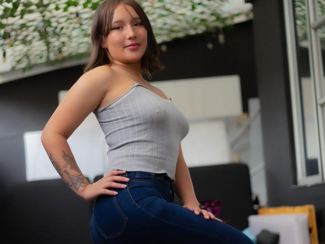 ColetteJohnson with medium brown hair, gray tank top, and tight blue jeans, showing off her tits and ass, hand on hip, tattoo on left arm, smiling at camera.