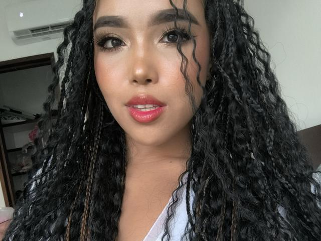 Heyly, con el pelo negro rizado largo, está chupando sus labios rosados brillantes, mirando a la cámara, usando el maquillaje pesado y un la camisa blanca.