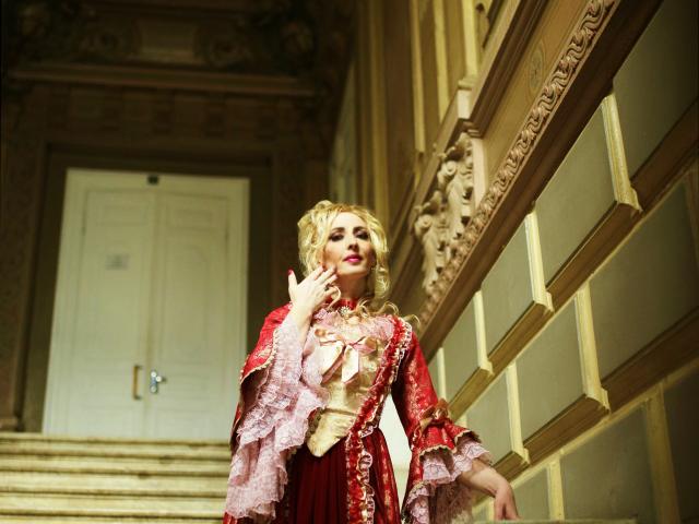Blonde EroLayer with curled hair in a red and gold, frilled, fantasy-style dress, standing on ornate stairs, hand raised to her face, looking upward.