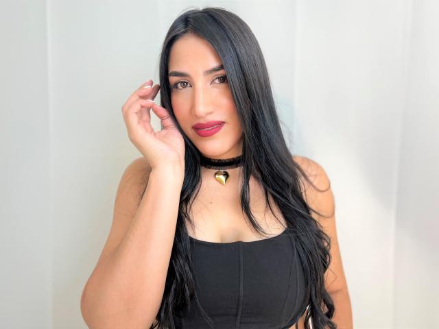 Alana Harris, con lunghi capelli neri, indossa una collana nera con un cuore dorato, un top nero con spalline, mentre tocca i capelli, ha il rossetto rosso, guarda la fotocamera, mostrando il seno..