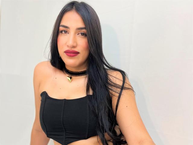 AlanaHarris with long black hair, wearing a black strapless top, choker with gold triangle, red lipstick, and brown eyes, showing her tits.