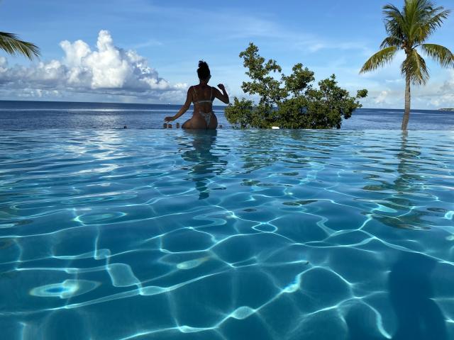 GalaMalkova in a blue bikini, standing waist-deep in a clear pool, holding a drink, with a small island and palm trees in the background, under a bright blue sky.
