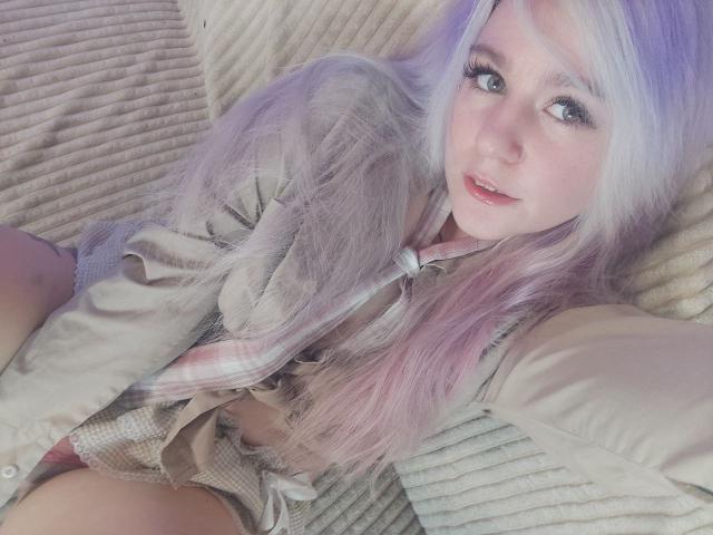 WhiteKrol with long, pastel purple and pink hair, wearing a plaid shirt and shorts, lying on a textured beige surface, looking at the camera with a soft smile.