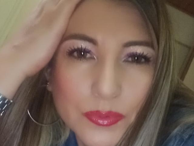 JolieMatureI's face, with her hand touching her blonde hair, showing pink eyeshadow, long eyelashes, red glossy lips, and a silver bracelet. Her hoop earrings are visible.