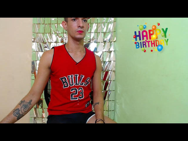 NathanTwentyTwo, slim with tattoos, wearing a red "Builds 23" basketball jersey, black shorts, and pink lipstick, sitting against a shiny green wall. Birthday decorations on the right.