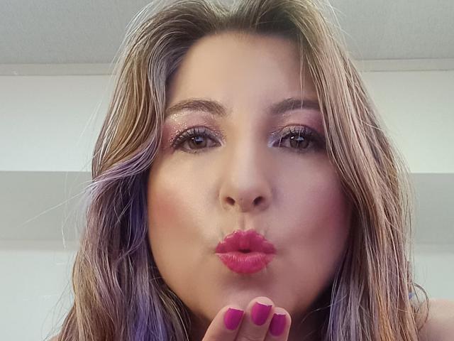 JolieMatureI has pink lipstick, shiny purple eyes, and pink-painted nails, pouting her glossy pink lips with her finger, brown wavy hair with purple highlights, smiling.