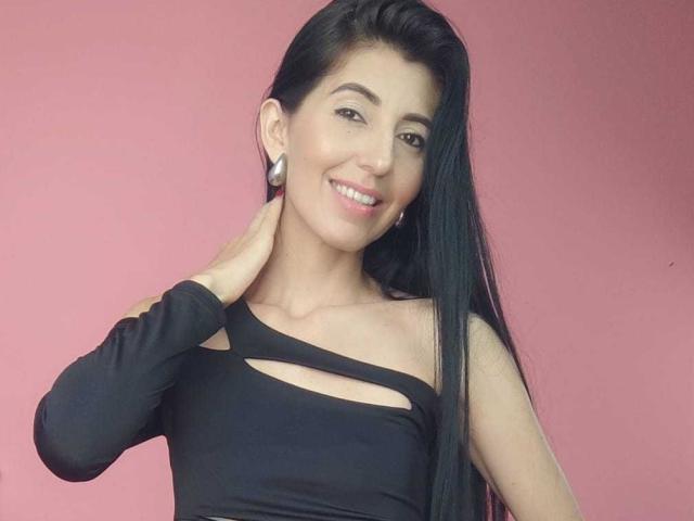 SweetSheilaM with long black hair, smiling, wearing a black one-shoulder top with cutout, touching her neck, showing small white earrings, pink background, and light skin.