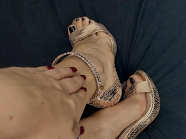 LizzieRossa's foot with snake-skin heels, black-painted toenails, and a hand with red nails resting on her ankle. Her skin is fair, and the background is dark.