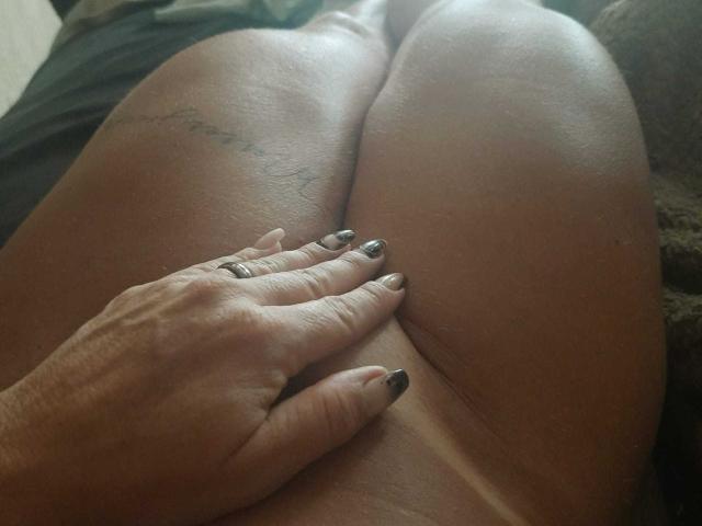 MistressBarbaraO's thighs, her hand with black nail polish resting on her crotch, her tattoo visible on her upper thigh. Her skin is tan, and she's nude.