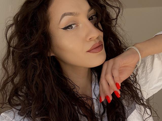 VievienneClemente with wavy dark hair, bold eyeliner, red lipstick, red nails, white shirt, and silver bracelet, looking confidently at the camera. Her hand rests near her face.