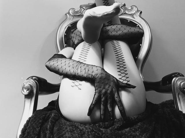 IzzysMisss is sitting on a fancy chair, legs up, wearing fishnet gloves, and holding her butt with one hand. Her ass is prominently displayed. Black and white photo.