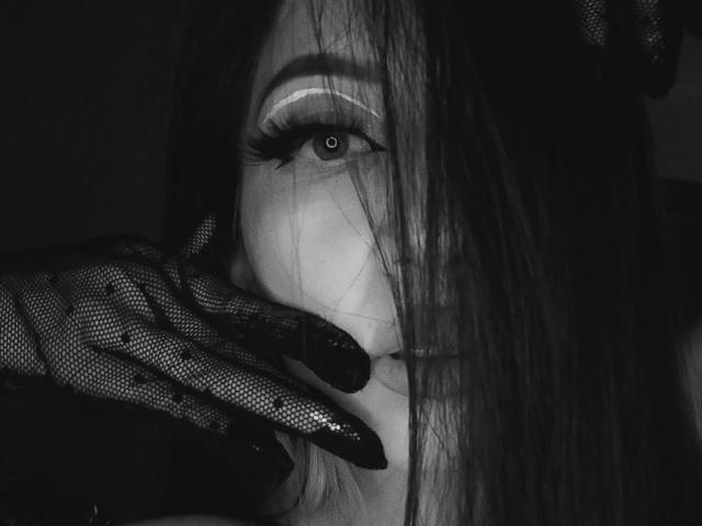 IzzysMisss with long hair, black mesh glove, and thick eyelashes, touching her glossy lips with a finger, in a dark, moody, black-and-white photo.