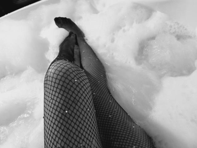 IzzysMisss' legs in black fishnet stockings, partially submerged in white bubbly water, with her feet crossed at the toes. Black and white photo, focus on legs.
