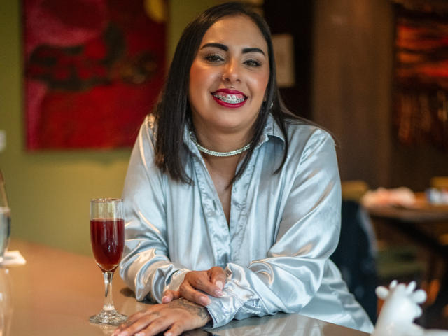 Genexis with dark hair, red lipstick, and shiny silver shirt, smiling, hands on table, red wine glass in front, chandelier necklace, in a cozy bar.